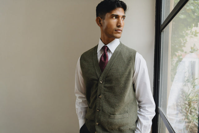 Man stood looking out of the window wearing a green knitted vest with shirt and tie