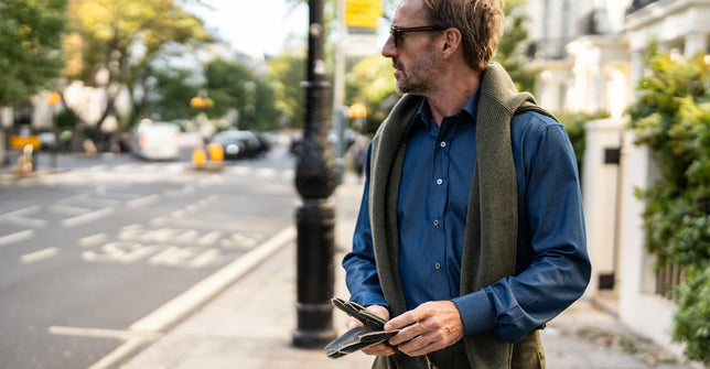 Denim shirt with seed stich jumper thrown over the shoulders, holding a pair of leather gloves in the street