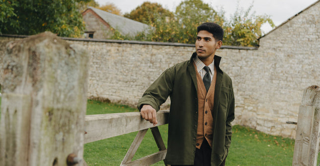 Man stood in field by wooden gate wearing knitwear and safari jacket