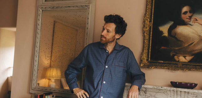 Male model sat in armchair looking out onto the garden wearing sky blue Budd pyjamas