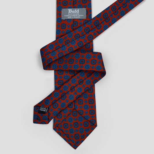 A folded necktie with a red base and a blue paisley pattern, featuring a visible 'Budd' brand label.