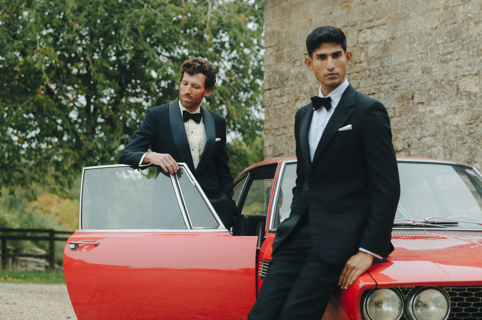 Two men in formal attire standing next to a red car outdoors.