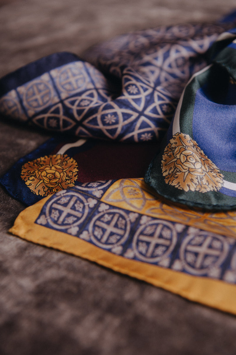 A close-up of a patterned silk scarf draped on a textured brown surface. The scarf features navy blue and gold traditional Chinese motifs, a gold embroidered medallion, and a green and blue corner detail.