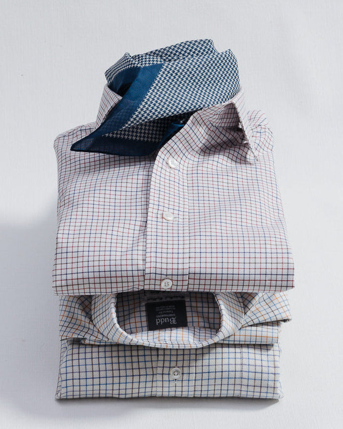 A stack of three neatly folded dress shirts with a small checkered pattern, with a blue striped necktie resting on top, set against a plain white background.