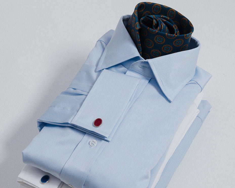 A neatly folded light blue dress shirt stack on a light gray background, topped with a rolled navy patterned tie and accented with red and blue cufflinks.