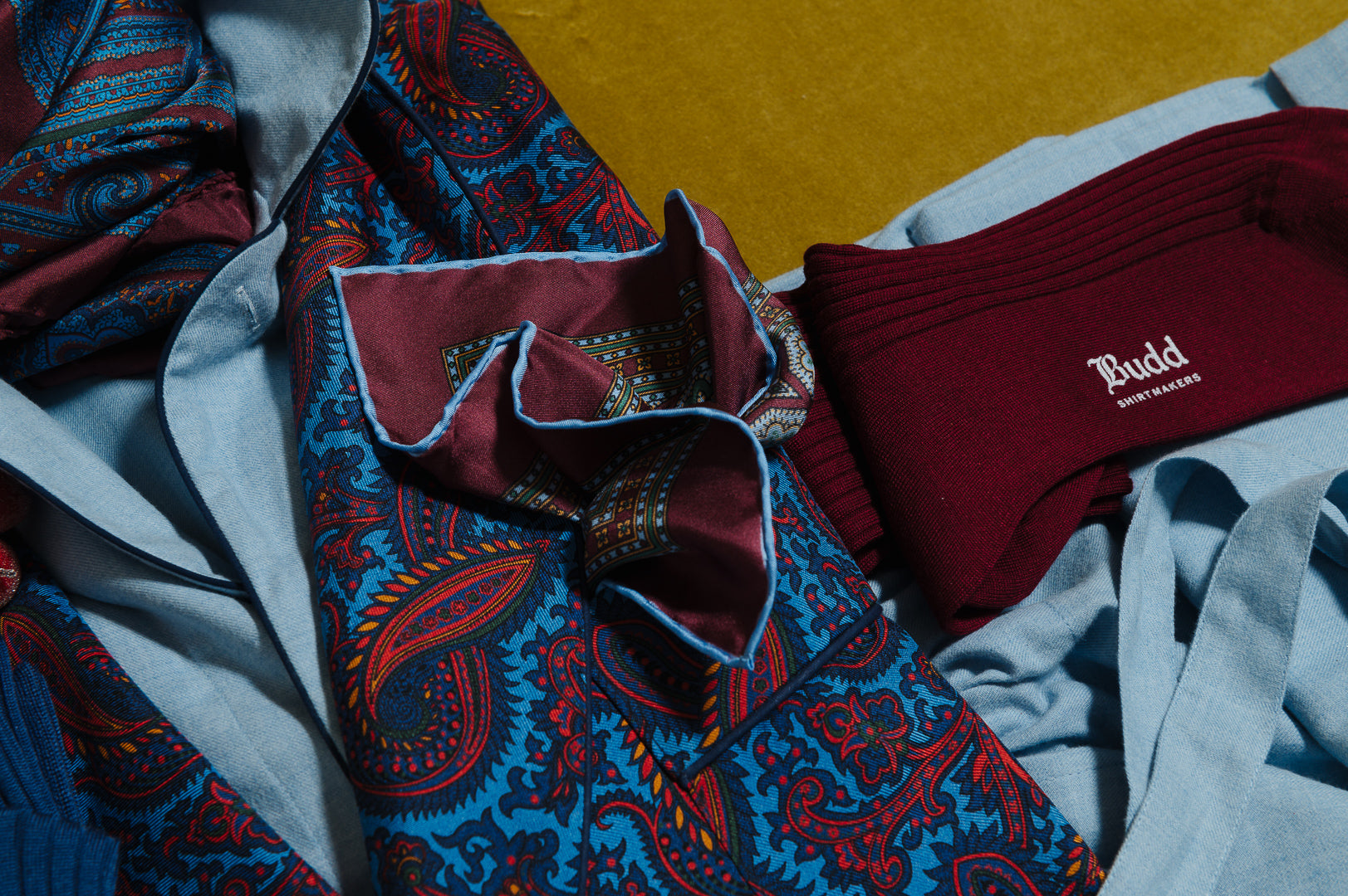 A styled flat-lay of men's fashion items including a blue and red paisley patterned silk tie, a burgundy ribbed knit neck warmer with 'Bludd' branding, and light blue dress shirts on a mustard yellow background.