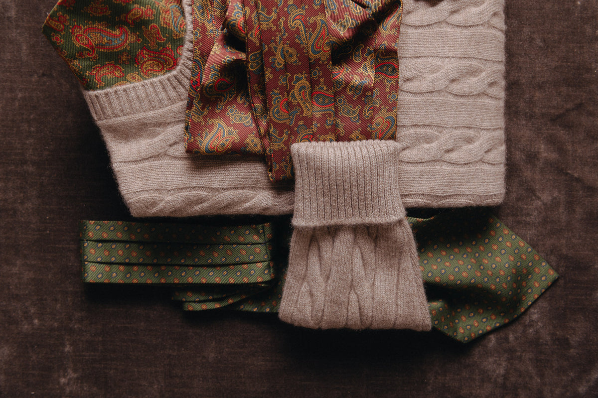A styled flat-lay of folded knit and patterned clothing on a dark brown textured surface, including cream cable-knit fabric, green paisley and polka-dot textiles, and red paisley fabric.