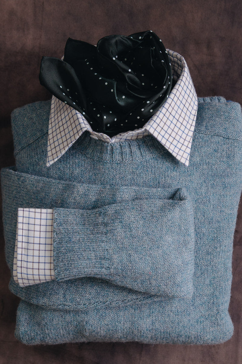 A folded gray knit sweater layered over a white and blue checkered dress shirt, with a black patterned neck scarf, arranged on a dark brown textured background.