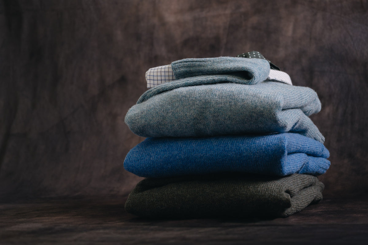 A stack of four folded knit sweaters in muted blue, teal, olive, and charcoal gray tones, arranged on a dark wooden surface against a dark textured backdrop.