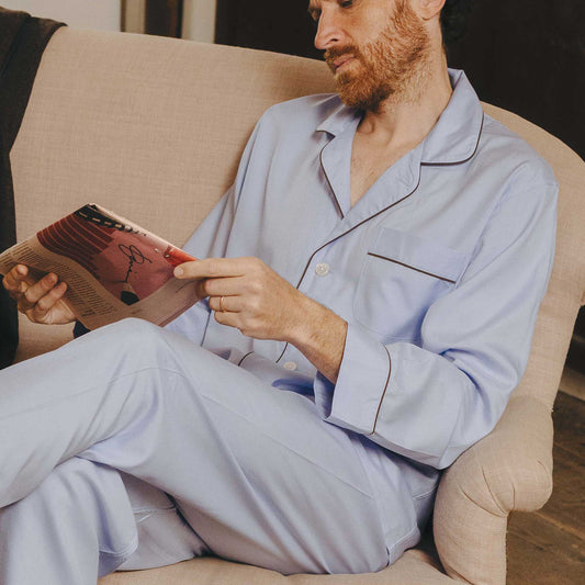 Man in light blue pajamas reading a book on a beige couch.