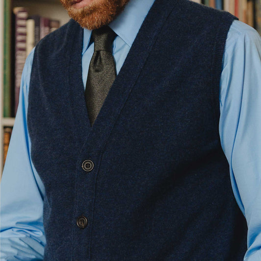 Man wearing a dark blue vest over a light blue shirt with a patterned tie, standing in front of a bookshelf.