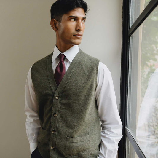 Man wearing a green vest, white shirt, and maroon tie standing by a window.