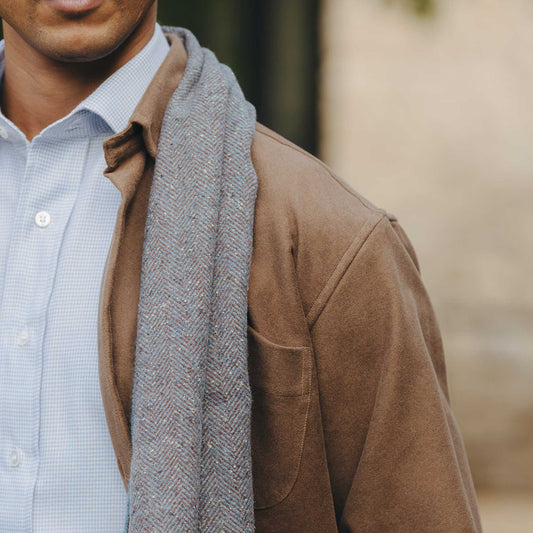 Man wearing a brown jacket, light blue shirt, and gray scarf with a blurred background