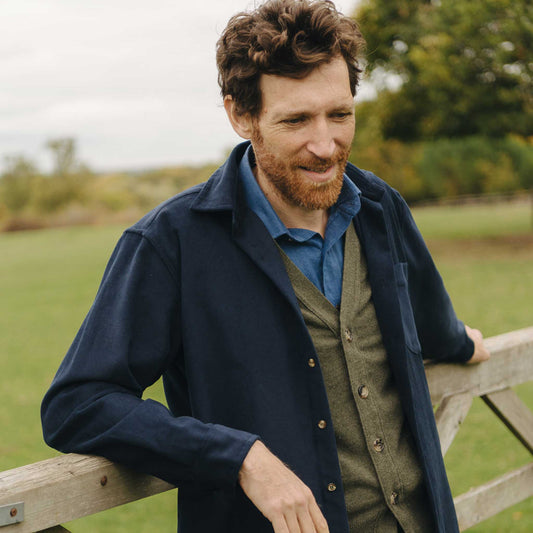 Man wearing a navy coat and green vest standing by a wooden fence in a field.