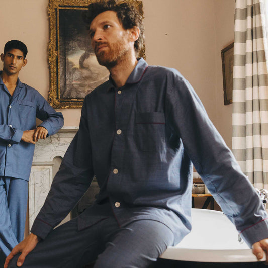 Man wearing a navy blue pyjama set sitting on a bed in a room with a painting and checkered curtain.