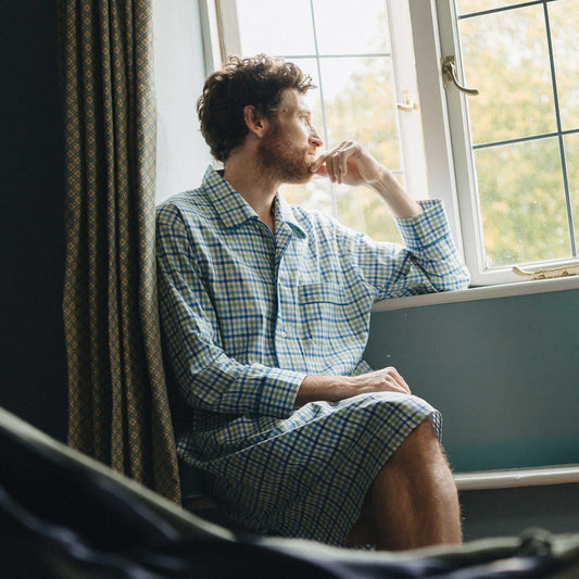 Man in a blue checked nightshirt sitting by a window with curtains.