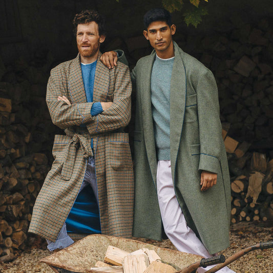 Male model shot of tweed dressing gown in green with checked pyjama shirt underneath stood outside in log store with another man wearing a checked gown