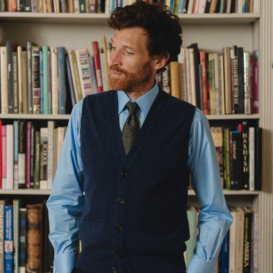 Man in a blue shirt and dark vest standing in front of a bookshelf