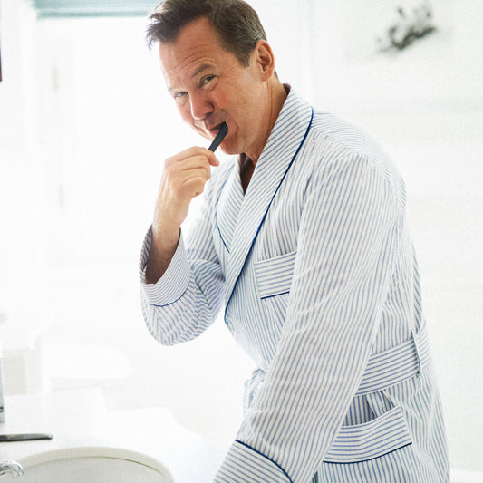 A man wearing a white and navy blue vertically striped bathrobe stands in a bright bathroom, holding a toothbrush near his mouth.