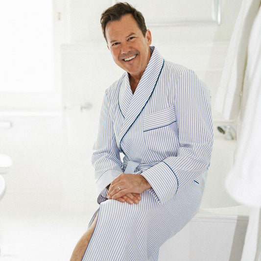 A smiling man wearing a light blue and white vertically striped bathrobe, seated in a bright, white bathroom setting.