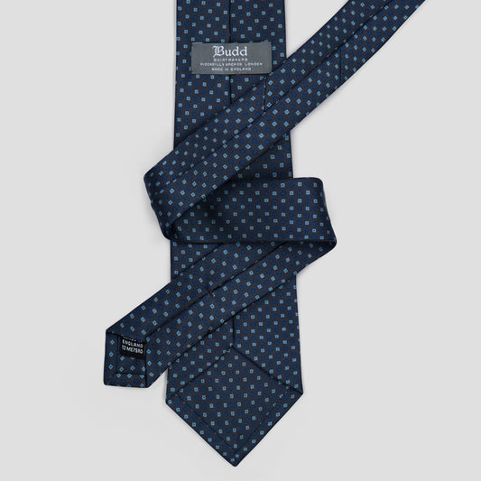 A navy blue silk tie with a small repeating geometric pattern in light blue and white, neatly folded and displayed on a light gray background. The brand label 'Budd' is visible on the inner neck band.
