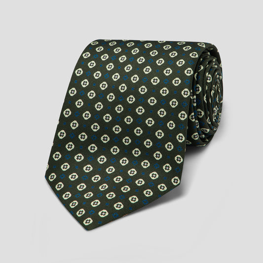 A rolled necktie with a dark olive green base, featuring a repeating pattern of white and light blue geometric medallions, set against a neutral light gray background.