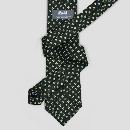 A dark green necktie with a repeating cream and gold circular pattern, laid flat on a light gray background. The brand label 'Budd' is visible near the collar.