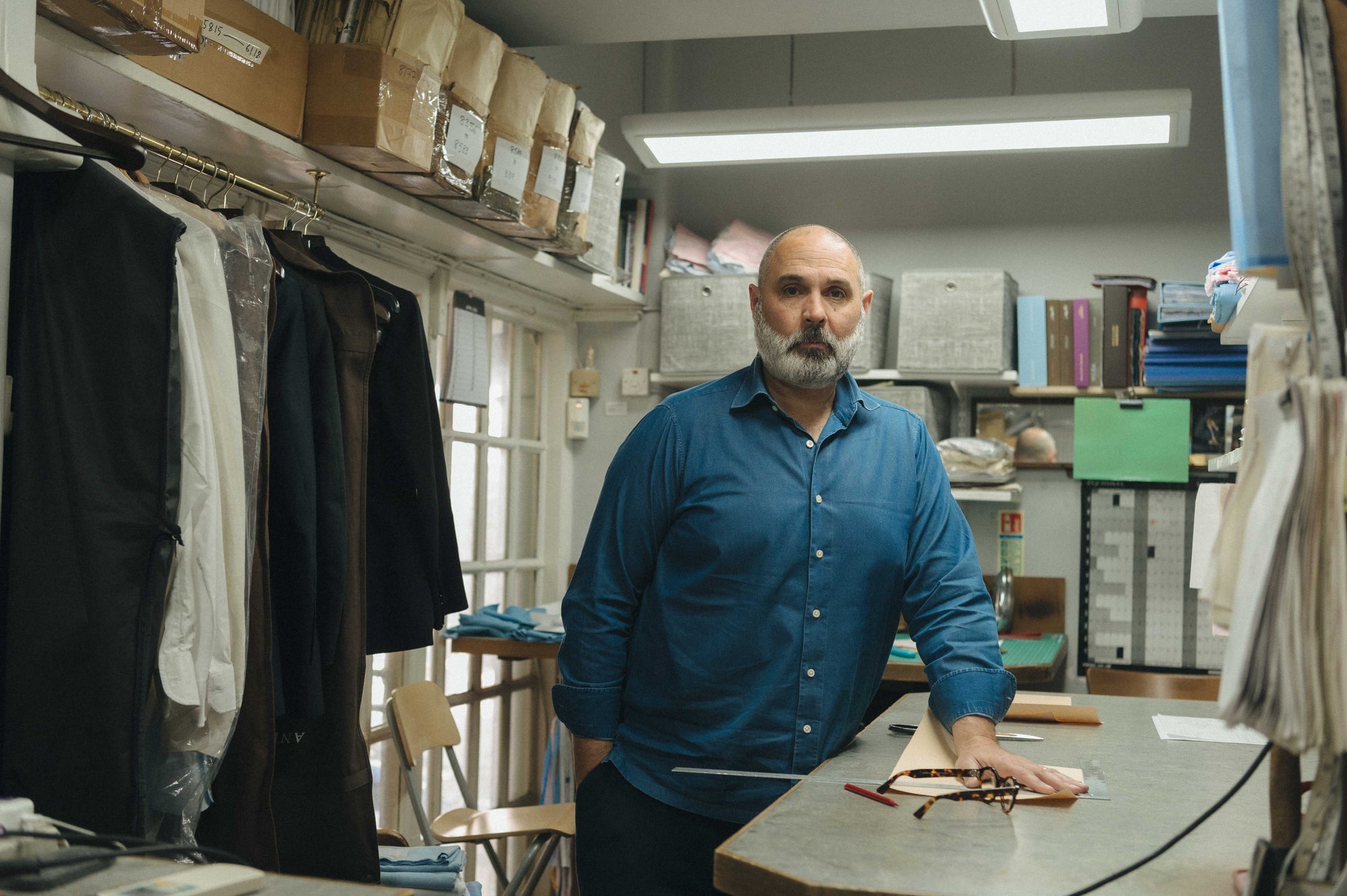 Man in a blue shirt standing in a workshop or storage room with shelves and clothing items.