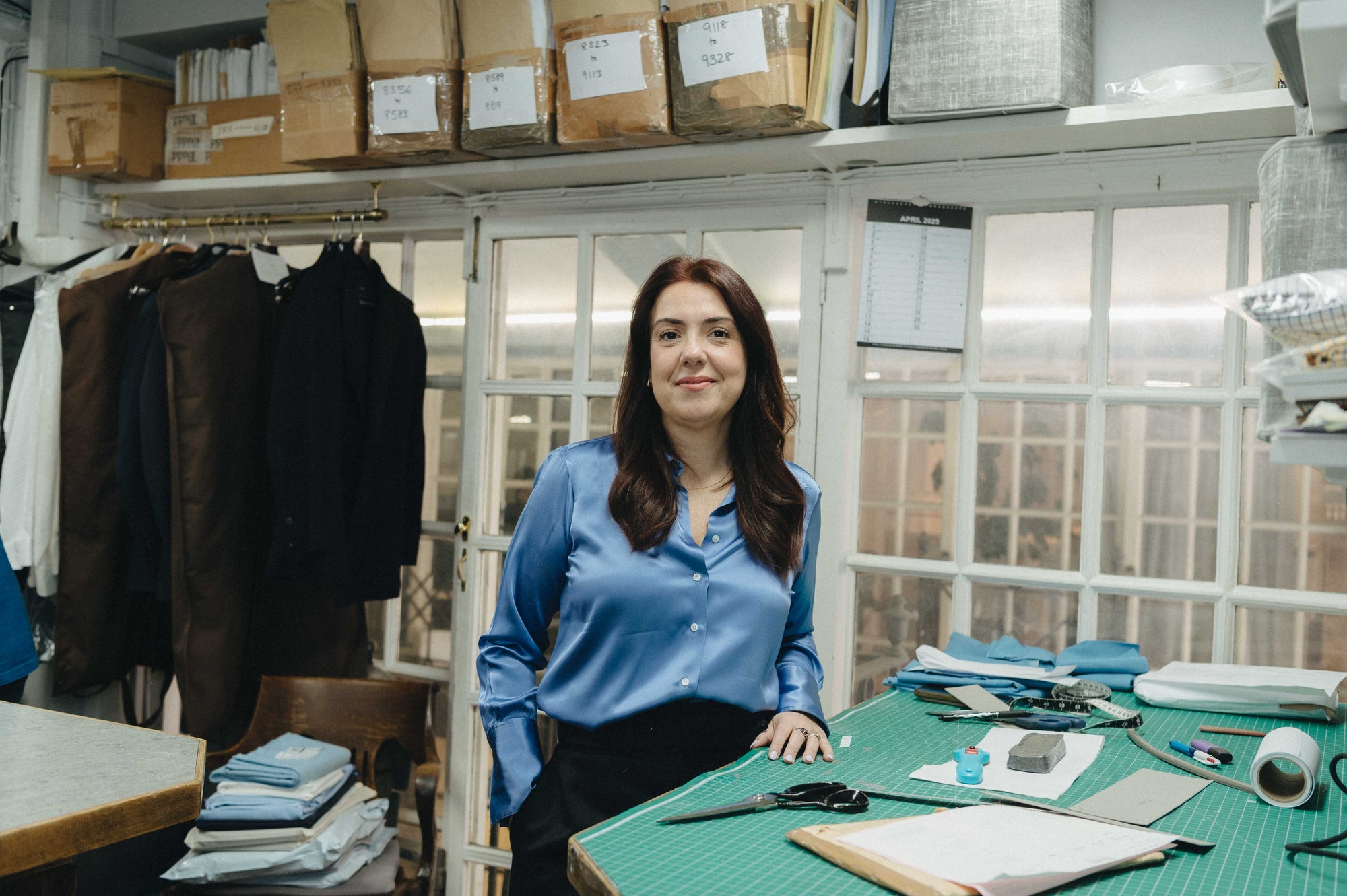 Woman standing in a clothing store with racks of clothes and a table with fabric and tools.