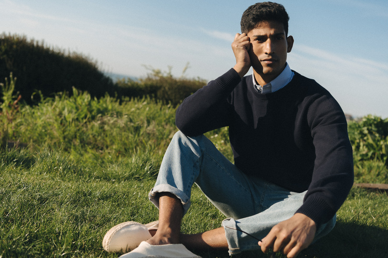 Man sat on grass by coast wearing a navy jumper and jeans