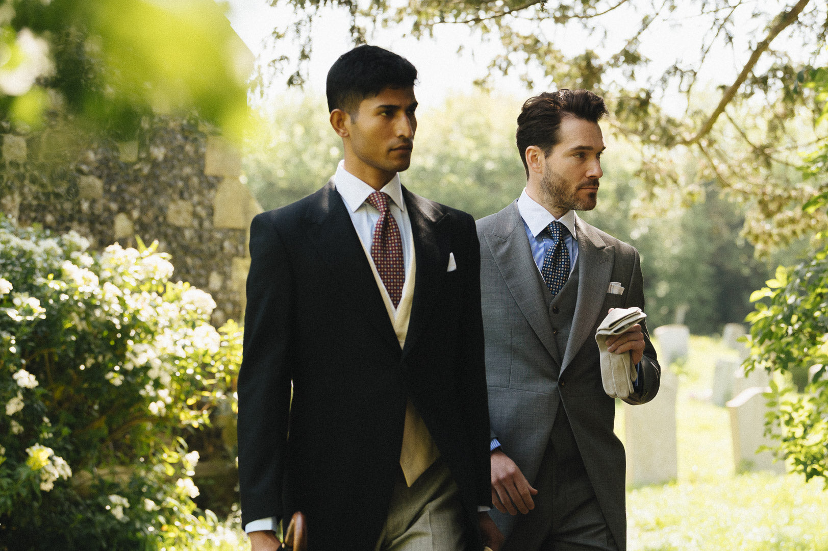 Two men in formal suits walking outdoors with greenery in the background