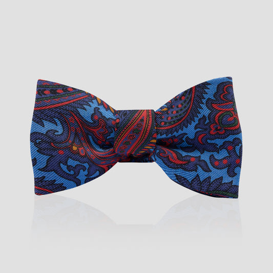 A pre-tied bow tie featuring a detailed paisley pattern in shades of blue, red, and black, presented on a light gray reflective background.