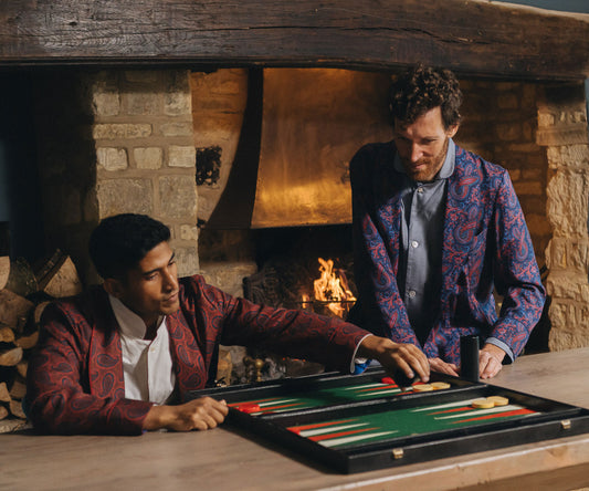Two men playing backgammon in a cozy room with a fireplace wearing paisley silk dressing gowns