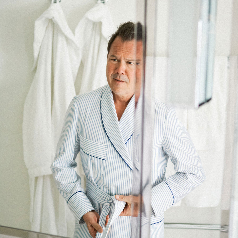 A man wearing a light blue and white vertically striped bathrobe stands in front of a mirror, with white robes hanging in the background.
