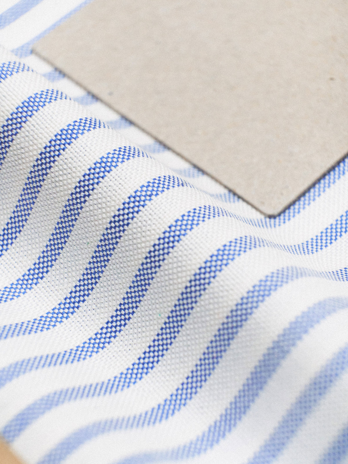 Close-up of a white woven fabric with repeating blue wavy stripes and a plain beige square sample placed on top.