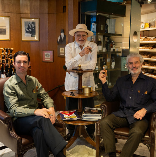 Three men sitting in a room with wooden paneling and shelves smoking cigars