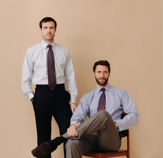 Two male models wearing business shirts with one stood up and one sat on chair against plain peach background