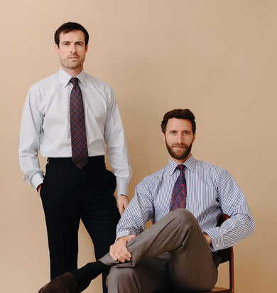 Two male models wearing business shirts with one stood up and one sat on chair against plain peach background