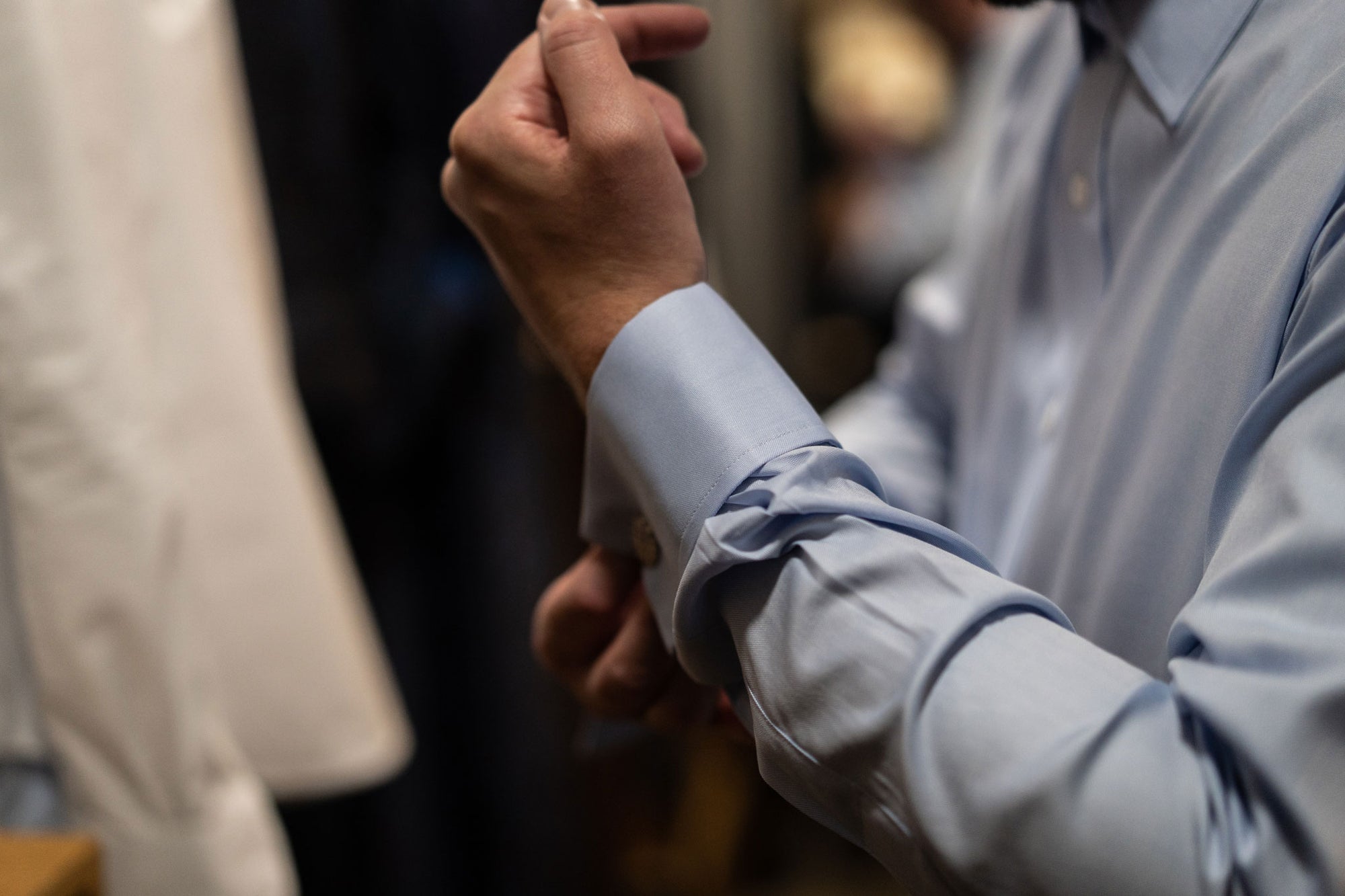 Person wearing a light blue shirt with rolled-up sleeves, standing in a blurred indoor setting.