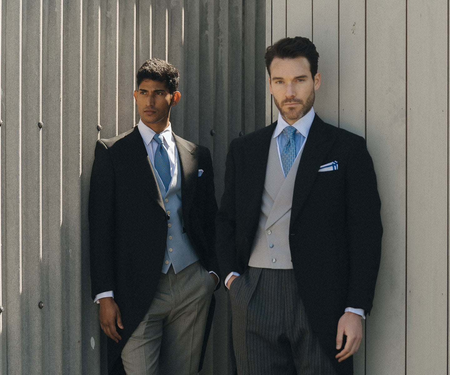 Two men dressed in wedding morning wear stood against grey building outside