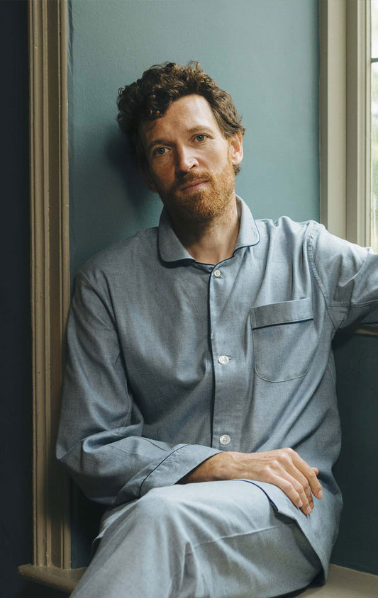 Man leaning against a window with a view of greenery wearing blue pyjamas