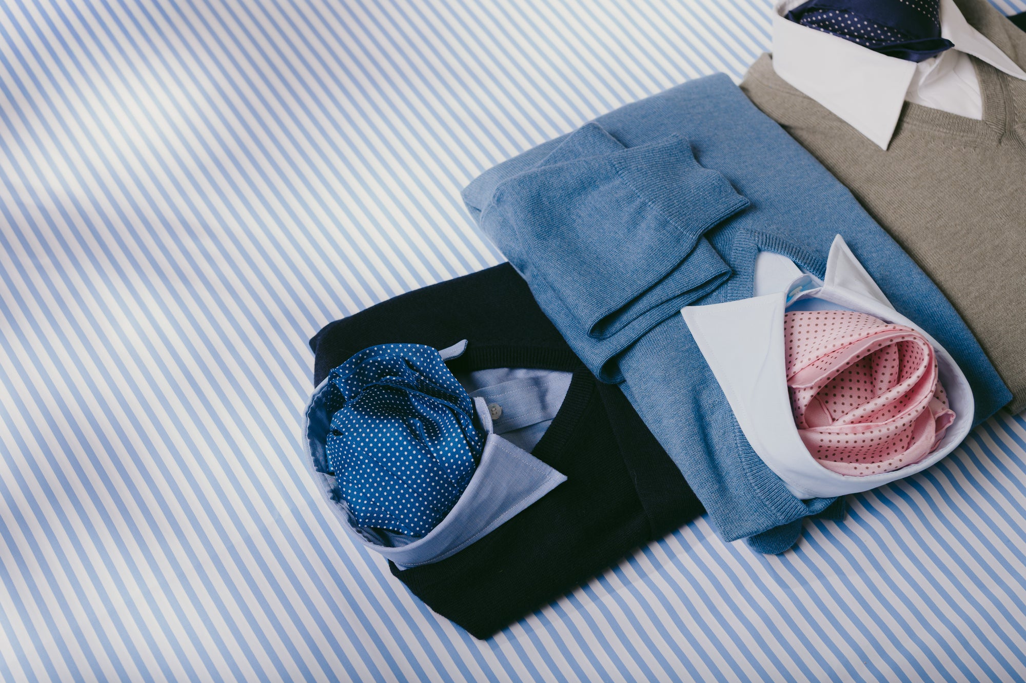Navy, beige and blue jumpers laid with shirts on blue stripe background