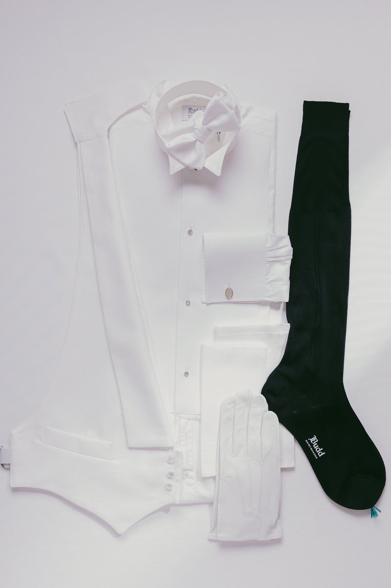 A flat-lay of a white formal outfit including a long white shirt, white vest, white bow tie, white pocket square, and a single black sock, arranged on a white background.