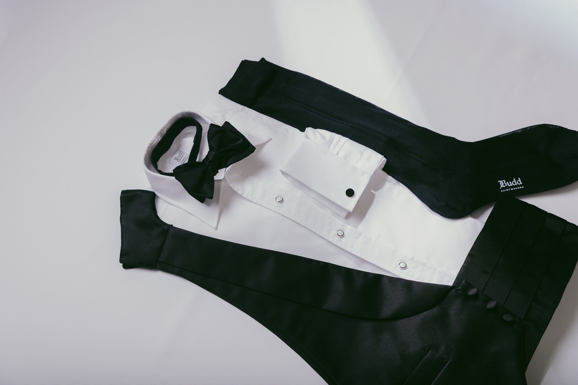 A formal men's outfit laid flat on a light background, including a white dress shirt with black bow tie, black vest, black trousers, and black socks. A small white 'Budd' brand tag is visible on one sock.