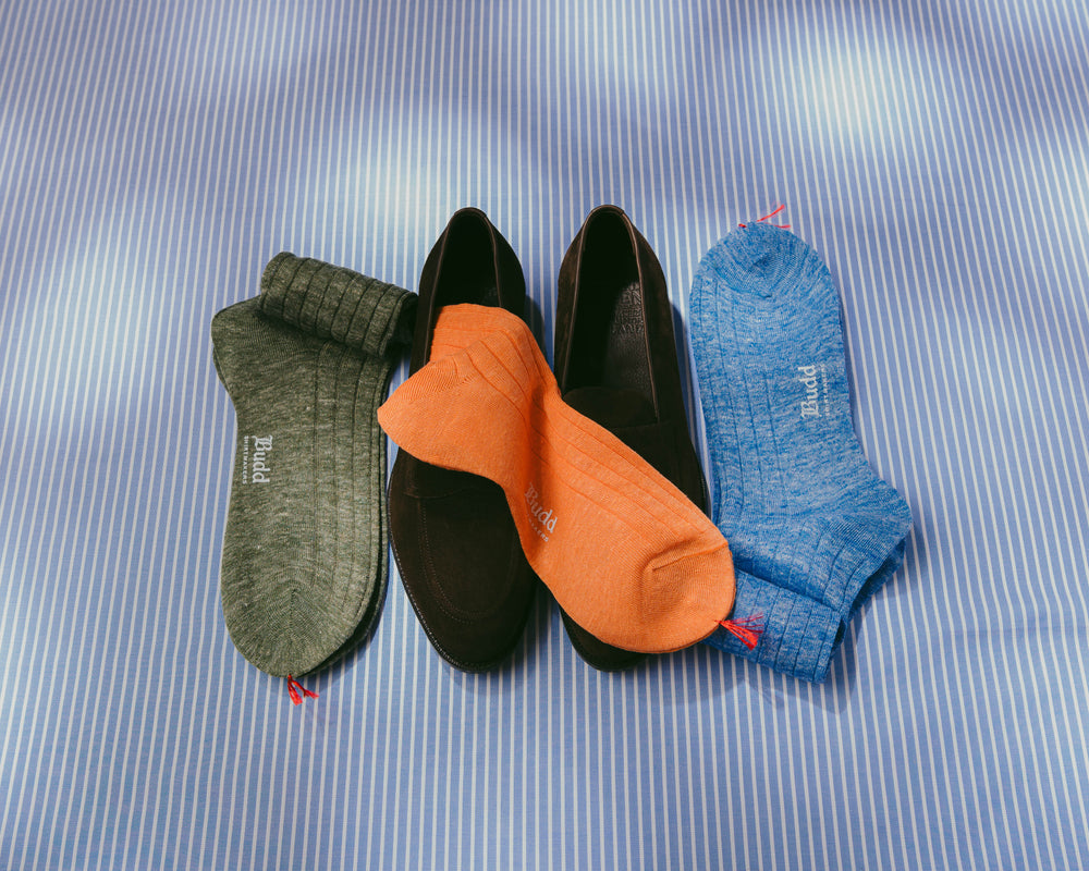 Four pairs of ankle socks arranged on a blue-and-white striped fabric background: olive green, black, orange, and heather blue. The socks are photographed from above with soft light and no person or real environment visible.